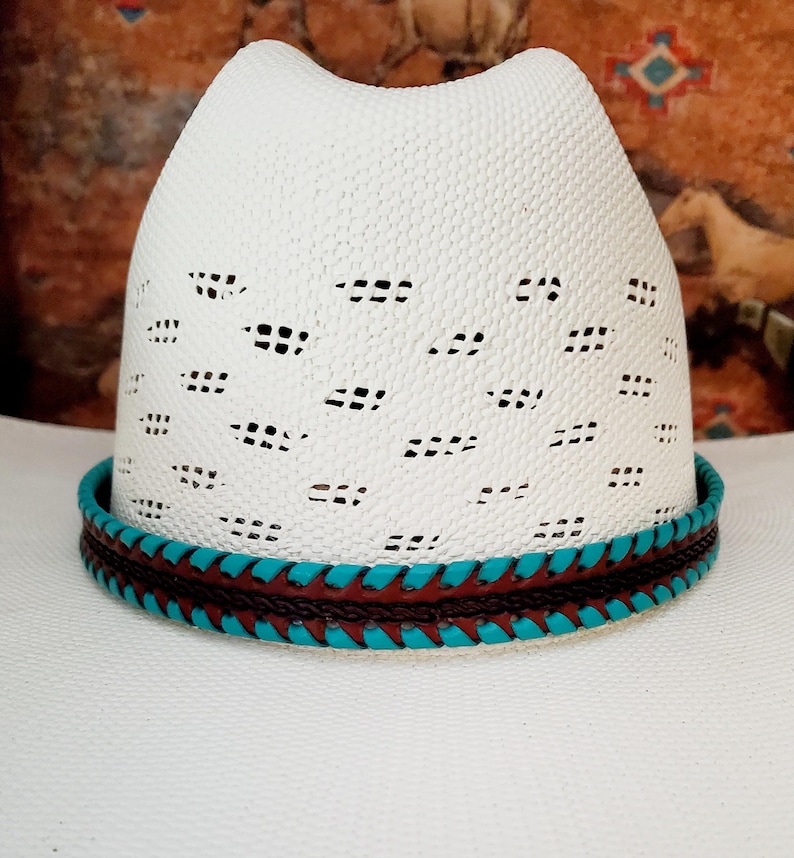 May include: A white cowboy hat featuring a ventilated crown with a repeating pattern of small rectangular cutouts. The base of the crown is encircled by a decorative hat band composed of braided dark brown leather and bright turquoise leather.