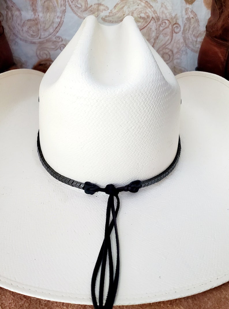 Pu&ograve; includere: Cappello da cowboy bianco con una fascia in pelle intrecciata nera e un cinturino per il mento in pelle nera.