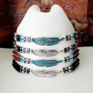 May include: Four hat bands wrapped around a white hat. Each band features a central metal feather charm in turquoise or silver, flanked by metallic cube beads, small round turquoise beads, and long cylindrical beads in black or brown.