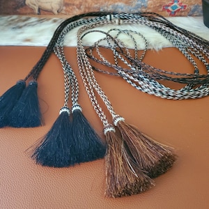 Puede incluir: Tres cordones de cuero trenzados con borlas. Los cordones son negros, marrones y blancos. Las borlas son negras, marrones y marrones y blancas.