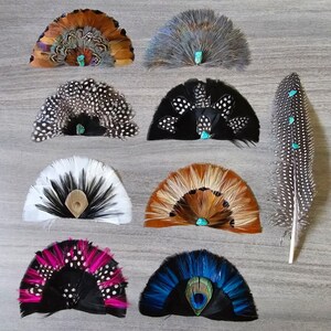 Puede incluir: Una colecci&oacute;n de abanicos de plumas hechos a mano en varios colores, incluyendo marr&oacute;n, negro, blanco y azul. Cada abanico presenta un acento turquesa. Tambi&eacute;n hay una pluma larga con acentos turquesa.
