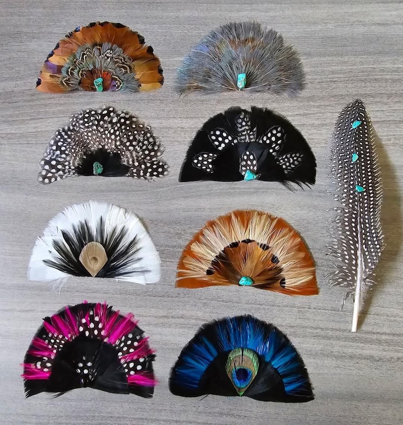 Puede incluir: Una colecci&oacute;n de abanicos de plumas hechos a mano en varios colores, incluyendo marr&oacute;n, negro, blanco y azul. Cada abanico presenta un acento turquesa. Tambi&eacute;n hay una pluma larga con acentos turquesa.