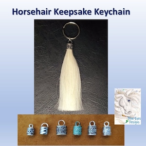 May include: White horsehair keychain with a silver key ring and a silver charm with an intricate design. The charm is attached to a white horsehair tassel. The keychain is displayed on a brown surface with other charms in various colors.