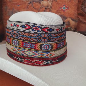 May include: A white cowboy hat featuring a stack of five distinct hat bands. Each band displays a unique woven geometric pattern in vibrant colors including red, blue, yellow, and black. The bands are layered horizontally around the crown.