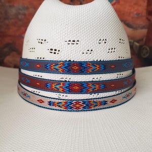 Op de afbeelding: Een witte fedora-hoed met een geperforeerde kroon en een decoratieve band. De band heeft een geometrisch patroon in tinten blauw, rood en geel. De hoed is een stijlvol accessoire.