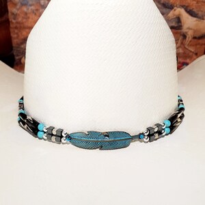 May include: A white cowboy hat with a turquoise and black beaded hat band featuring a metal feather charm.
