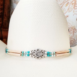 May include: A white cowboy hat with a decorative band. The band features light-coloured wooden beads, turquoise-coloured beads, and a silver-coloured floral centrepiece. The hat has a woven texture.
