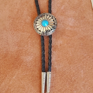 May include: A bolo tie featuring a round silver-toned metal centrepiece with a sunburst pattern and a central blue stone. The cord is made of black braided leather and terminates in two silver-toned metal tips with a ribbed texture design.