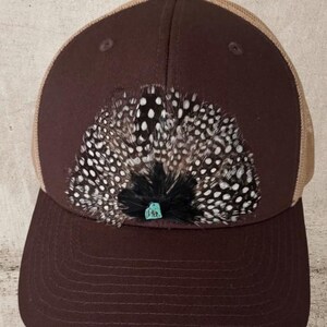 Puede incluir: Gorra de b&eacute;isbol marr&oacute;n con la parte trasera de malla color beige. La parte delantera de la gorra presenta una disposici&oacute;n decorativa de plumas con lunares blancos y un peque&ntilde;o acento de piedra turquesa. La gorra tiene una visera curva.