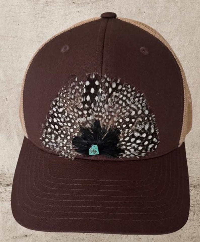 Puede incluir: Gorra de b&eacute;isbol marr&oacute;n con la parte trasera de malla color beige. La parte delantera de la gorra presenta una disposici&oacute;n decorativa de plumas con lunares blancos y un peque&ntilde;o acento de piedra turquesa. La gorra tiene una visera curva.