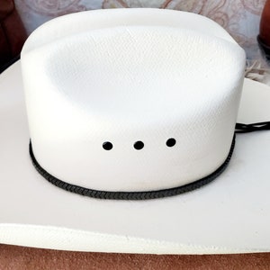 Pu&ograve; includere: Un cappello da cowboy bianco con una fascia intrecciata nera e tre occhielli neri. Il cappello &egrave; fatto di paglia e ha un'ampia tesa.