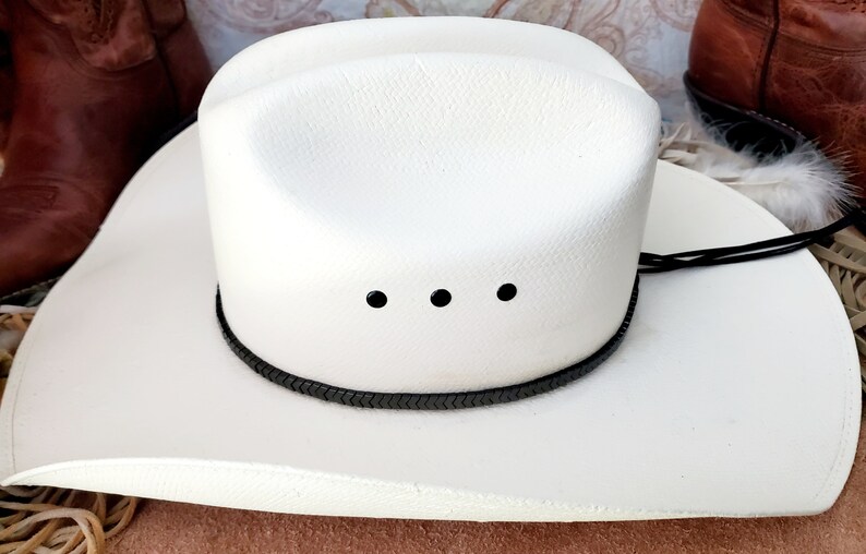 Pu&ograve; includere: Un cappello da cowboy bianco con una fascia intrecciata nera e tre occhielli neri. Il cappello &egrave; fatto di paglia e ha un'ampia tesa.