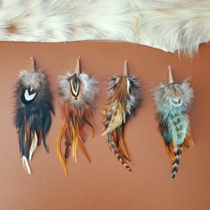 May include: Four decorative feather pendants with silver-coloured metal tops. The feathers are in shades of black, brown, orange, and teal. The pendants are arranged on a brown leather surface, with a white and tan fur-like material in the background.