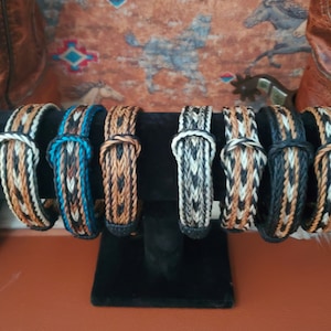 May include: A display of eight braided leather bracelets in various colors, including black, white, brown, and blue. The bracelets are displayed on a black stand.