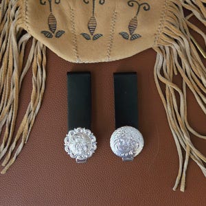 Puede incluir: Dos presillas de cinturón de cuero negro con hebillas decorativas plateadas. Las hebillas tienen un diseño floral ornamentado. Una bolsa de ante marrón claro con flecos está en el fondo, sobre una superficie de cuero marrón.