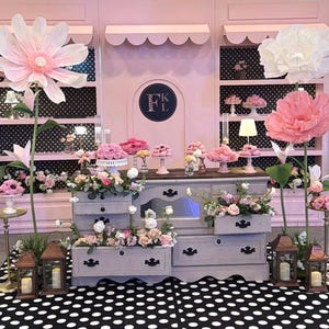 May include: A display featuring a light gray wooden dresser with open drawers filled with floral arrangements, flanked by two large paper flowers on tall stems. The background consists of pink shelving units with black and white polka dot backings.