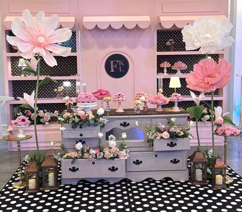 May include: A display featuring a light gray wooden dresser with open drawers filled with floral arrangements, flanked by two large paper flowers on tall stems. The background consists of pink shelving units with black and white polka dot backings.