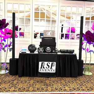 May include: A DJ setup on a black-skirted table featuring a black RSF EVENTS banner, two tall vertical speakers, a central console with a disco ball, and a sign reading DJ. Large decorative purple and pink flowers stand on both sides of the table.