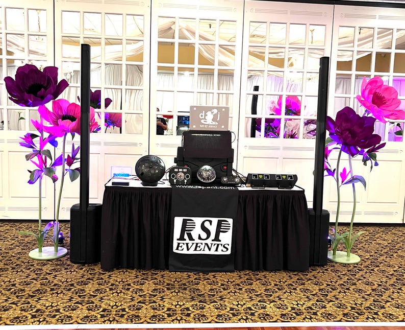 May include: A DJ setup on a black-skirted table featuring a black RSF EVENTS banner, two tall vertical speakers, a central console with a disco ball, and a sign reading DJ. Large decorative purple and pink flowers stand on both sides of the table.