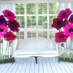 May include: A white tufted sofa with curved arms and dark wooden legs, flanked by two clusters of large, oversized artificial flowers in shades of magenta, deep pink, and purple, each mounted on green stems with grass-like bases.