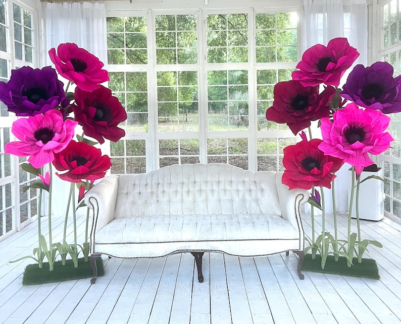 May include: A white tufted sofa with curved arms and dark wooden legs, flanked by two clusters of large, oversized artificial flowers in shades of magenta, deep pink, and purple, each mounted on green stems with grass-like bases.