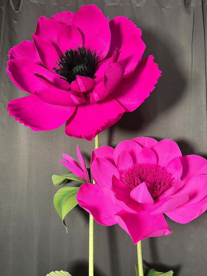 May include: Two large, vibrant fuchsia paper flowers with layered, ruffled petals and dark, textured centers. Each flower is attached to a slender, light green stem with small, pointed green leaves extending from the base of the blooms.