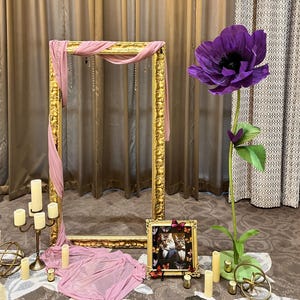 May include: A large ornate gold frame draped with dusty pink fabric, a tall artificial purple flower on a green base, a small gold frame containing a cat photograph, a gold candelabra with white candles, and several small gold decorative spheres.