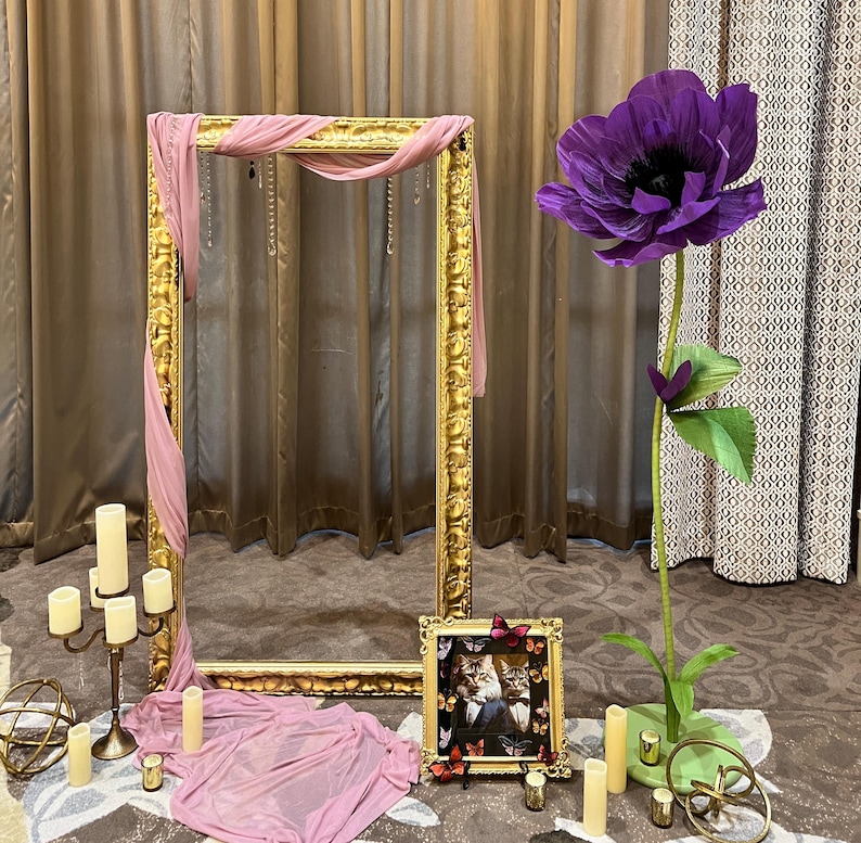 May include: A large ornate gold frame draped with dusty pink fabric, a tall artificial purple flower on a green base, a small gold frame containing a cat photograph, a gold candelabra with white candles, and several small gold decorative spheres.