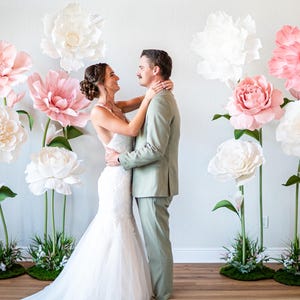 May include: A white wedding dress and a light green suit worn by two people standing together. The scene is decorated with several large, oversized paper flowers in shades of white and soft pink, each mounted on tall green stems with green bases.