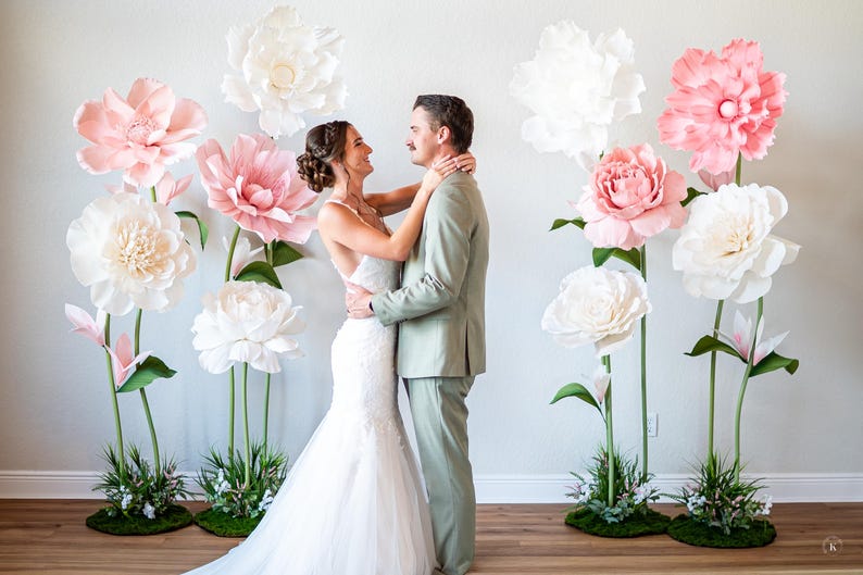 May include: A white wedding dress and a light green suit worn by two people standing together. The scene is decorated with several large, oversized paper flowers in shades of white and soft pink, each mounted on tall green stems with green bases.