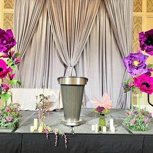 May include: A central gray podium stands on a stage flanked by two large floral arrangements featuring oversized magenta and purple paper flowers, green foliage, and small pink blossoms, with a gold candelabra and white candles nearby.