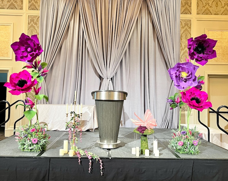 May include: A central gray podium stands on a stage flanked by two large floral arrangements featuring oversized magenta and purple paper flowers, green foliage, and small pink blossoms, with a gold candelabra and white candles nearby.