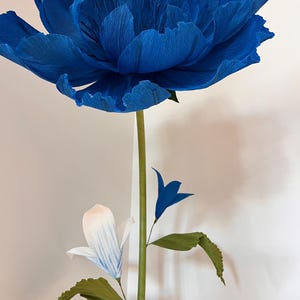 May include: A large, deep blue crepe paper flower with ruffled petals sits atop a long green stem. Below the main bloom, a smaller white flower with blue-tipped petals and a tiny blue bud are attached to the stem, along with two green leaves.