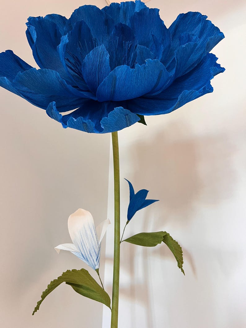 May include: A large, deep blue crepe paper flower with ruffled petals sits atop a long green stem. Below the main bloom, a smaller white flower with blue-tipped petals and a tiny blue bud are attached to the stem, along with two green leaves.