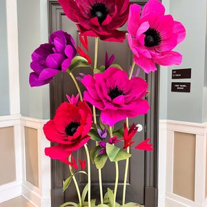 May include: A large decorative floral arrangement featuring oversized, vibrant flowers in shades of deep red, bright magenta, and purple. Each flower has a dark center and is supported by long, slender green stems with small green leaves.