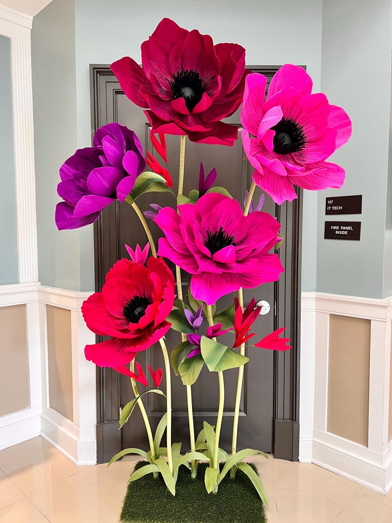 May include: A large decorative floral arrangement featuring oversized, vibrant flowers in shades of deep red, bright magenta, and purple. Each flower has a dark center and is supported by long, slender green stems with small green leaves.