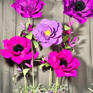 May include: A collection of large, handcrafted paper flowers in vibrant shades of magenta, fuchsia, and purple. The flowers feature dark centers, layered petals, and green stems, arranged with smaller floral accents and greenery at the base.