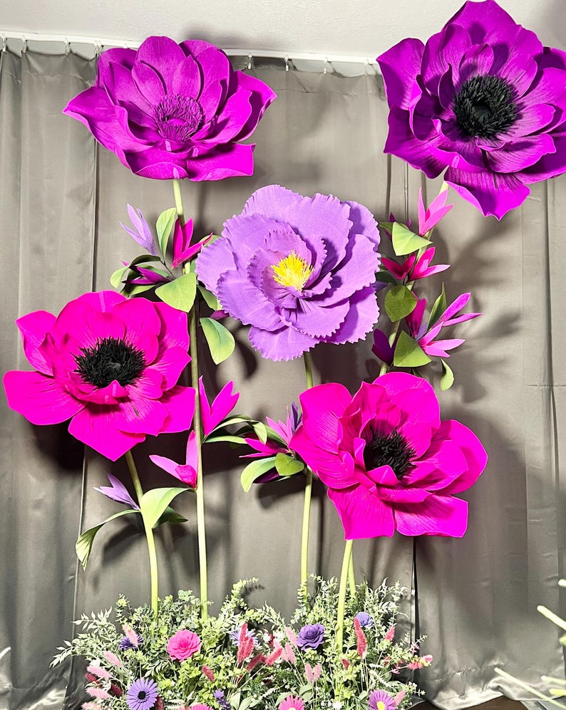 May include: A collection of large, handcrafted paper flowers in vibrant shades of magenta, fuchsia, and purple. The flowers feature dark centers, layered petals, and green stems, arranged with smaller floral accents and greenery at the base.