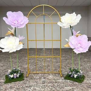 May include: A gold metal arched frame with a grid pattern, flanked by two floral arrangements. Each arrangement features large paper flowers in white and light purple, accented with gold leaves and small green plants on a mossy base.
