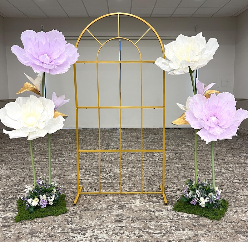 May include: A gold metal arched frame with a grid pattern, flanked by two floral arrangements. Each arrangement features large paper flowers in white and light purple, accented with gold leaves and small green plants on a mossy base.