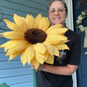 Puede incluir: Un gran girasol de papel amarillo con un centro marrón es sostenido por una persona que lleva una camisa negra. El girasol está hecho con pétalos de papel arrugados.
