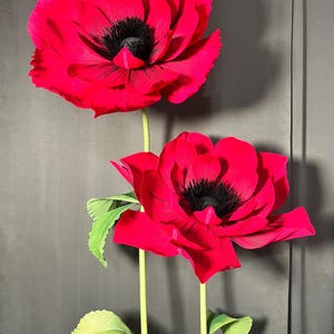 May include: Two large, handcrafted red poppy flowers with layered, textured petals and prominent black centers. Each flower is attached to a long, slender green stem featuring small, serrated green leaves extending from the sides of the stalks.