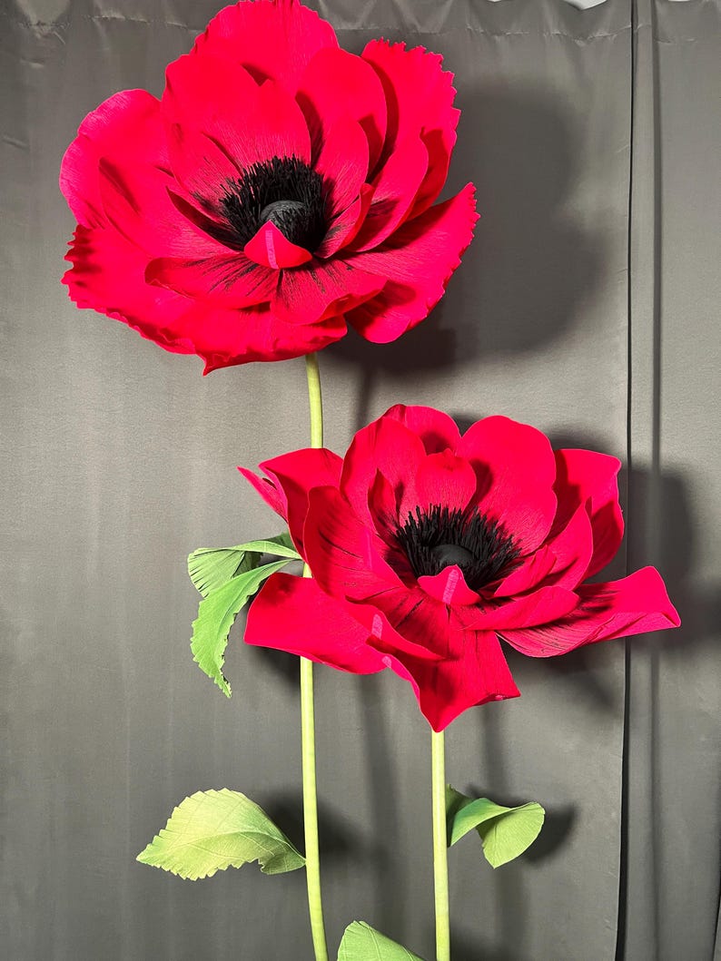 May include: Two large, handcrafted red poppy flowers with layered, textured petals and prominent black centers. Each flower is attached to a long, slender green stem featuring small, serrated green leaves extending from the sides of the stalks.