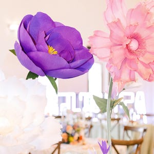 May include: Large handcrafted paper flowers featuring a deep purple bloom with a yellow center, a soft pink ruffled blossom, and a white flower. Each flower is attached to a thin green stem with green leaves, creating a vibrant floral display.