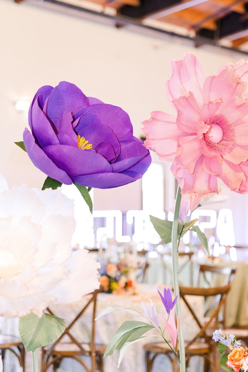 May include: Large handcrafted paper flowers featuring a deep purple bloom with a yellow center, a soft pink ruffled blossom, and a white flower. Each flower is attached to a thin green stem with green leaves, creating a vibrant floral display.