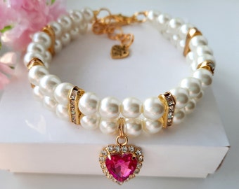 Puede incluir: Un collar de perro de doble fila de perlas con un dije de corazón dorado y un colgante de corazón de cristal rosa.