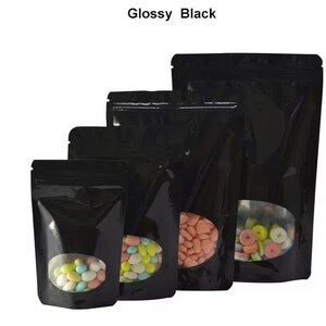 May include: Four black stand-up pouches with clear oval windows, the smallest pouch is filled with colorful candies, the next size up is filled with pastel candies, the next size up is filled with pink heart-shaped candies, and the largest pouch is empty.  Glossy Black.