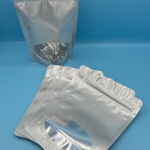 May include: A stack of white stand-up pouches with a clear oval window. The pouches have a zipper closure.