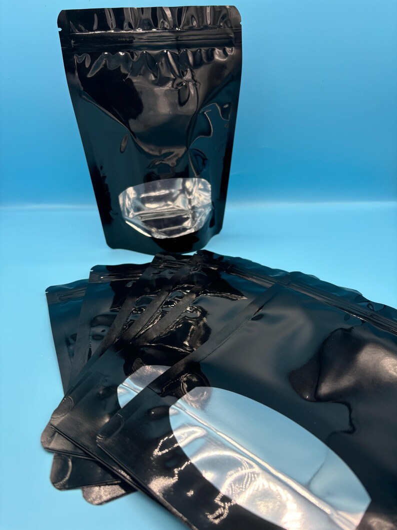 May include: A stack of black stand-up pouches with a clear oval window. The pouches have a zipper closure and are made of a shiny, metallic material.