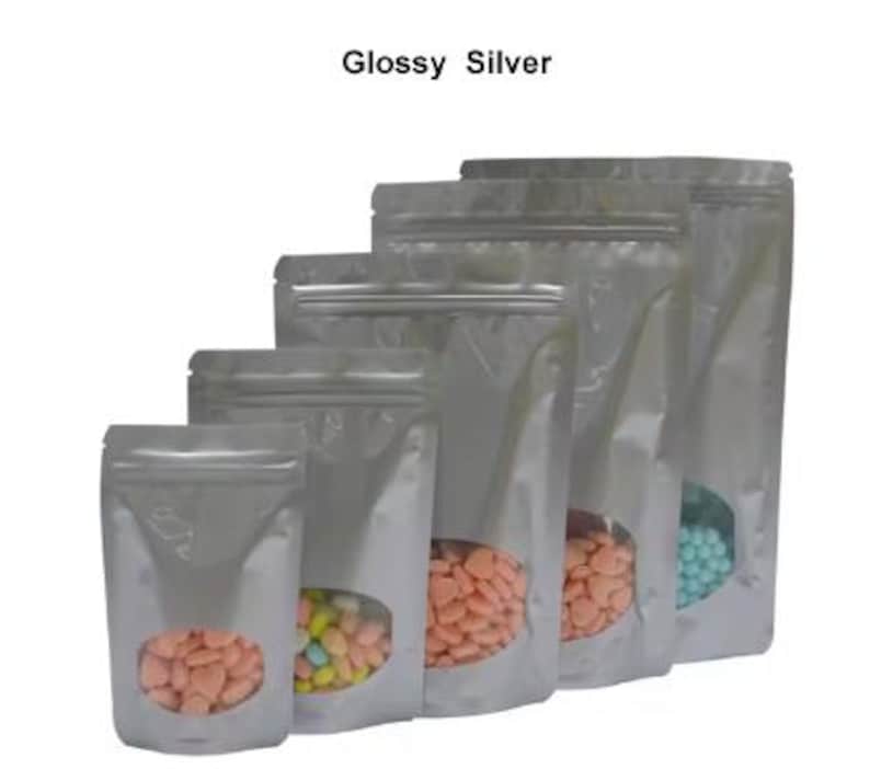 May include: Five silver stand-up pouches with clear viewing windows. The pouches are different sizes and contain colorful candies. The text "Glossy Silver" is at the top of the image.